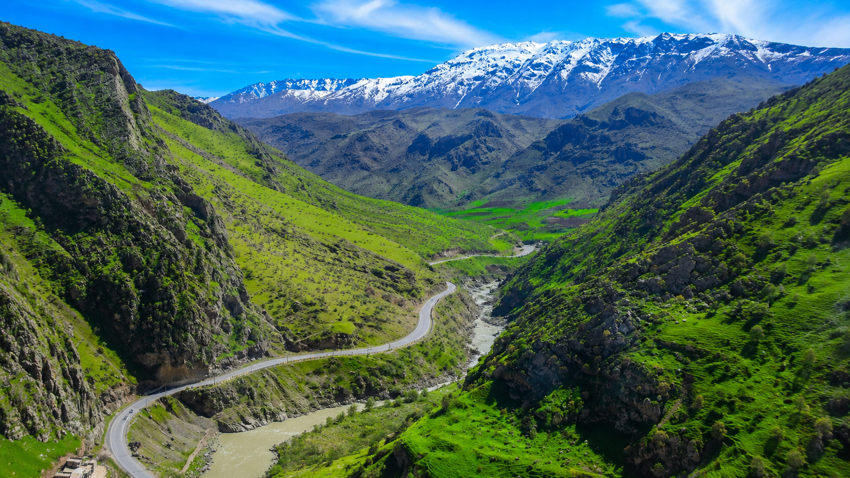 A road winding through a green valley beneath snow-capped mountains in the Kurdistan Region of Iraq